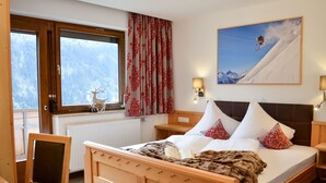 A wooden bed with two pillows, a nightstand, a lamp, a window with curtains, a radiator, a chair, and a picture of a skier on the wall. - Wechner by Interhome (Kappl)