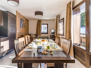 A dining area with a long wooden table set for breakfast, a kitchen with modern appliances, and a cozy seating area with a sofa and a TV. - Les Fermes Emiguy by Interhome (Les Gets)