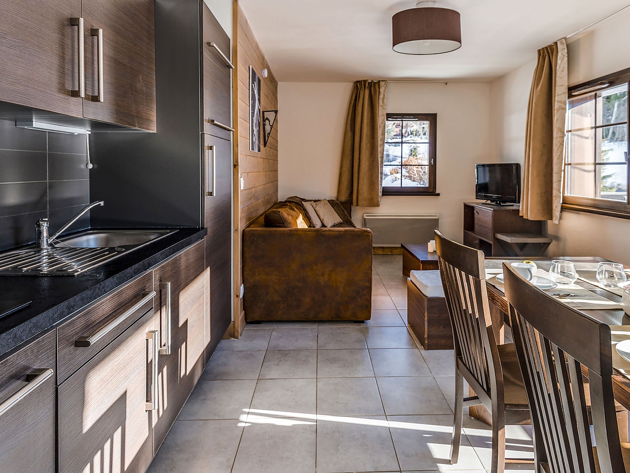 A modern kitchen with dark cabinetry, a black countertop, and a built-in oven. There is a dining area with a wooden table and chairs, and a sofa in the background.