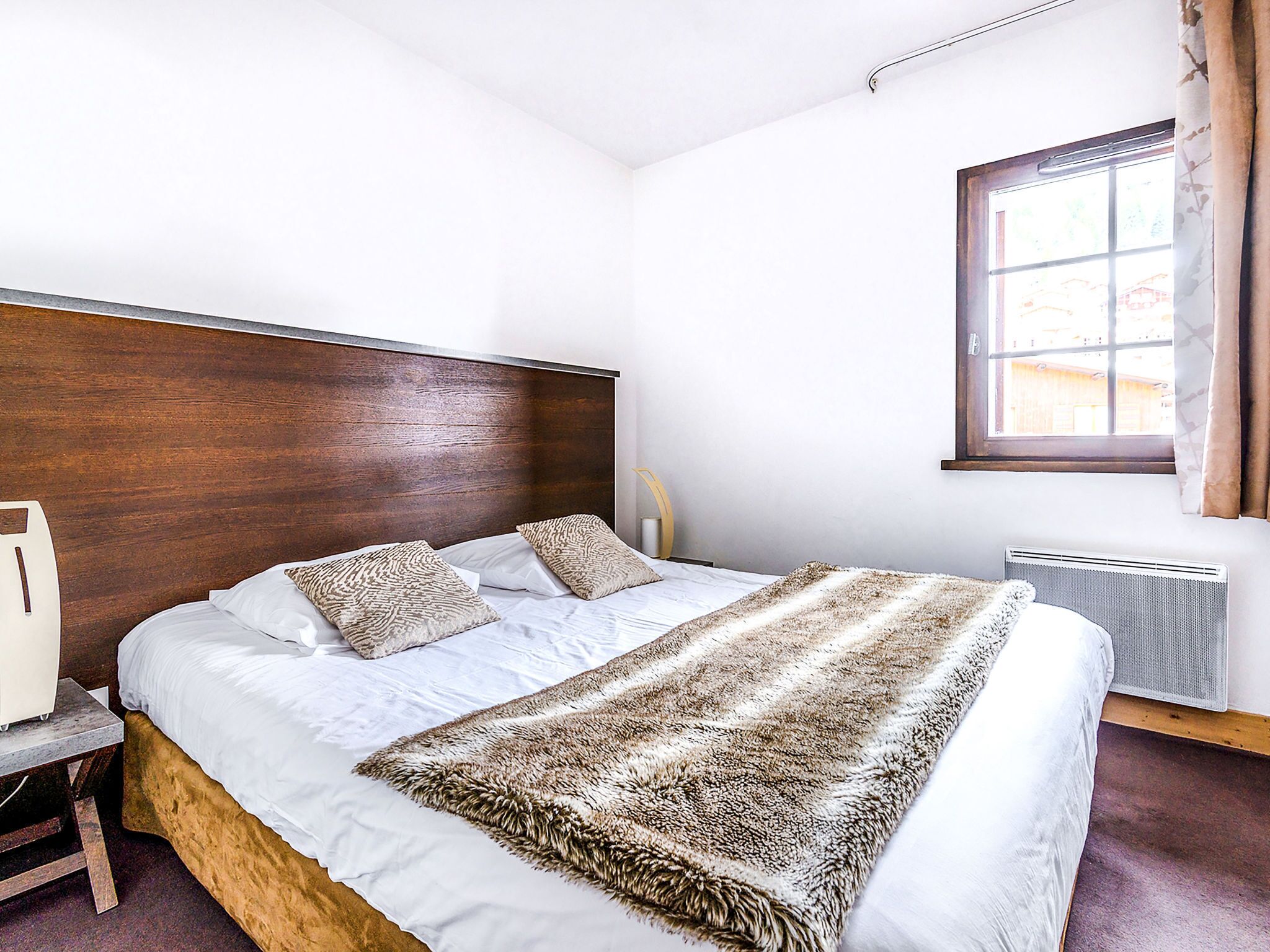 A bedroom with a wooden headboard, a bed with white linens and patterned pillows, a fur throw, a bedside table, and a window with curtains.