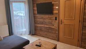A room with a wooden wall, a flat-screen TV mounted above a door, a sofa, a small wooden table with a remote control, and a window with curtains. - Apartment Köhle by Interhome (Pfunds-Samnaun)