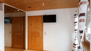 Quarto com piso de madeira, uma cama, televisão na parede e uma porta.