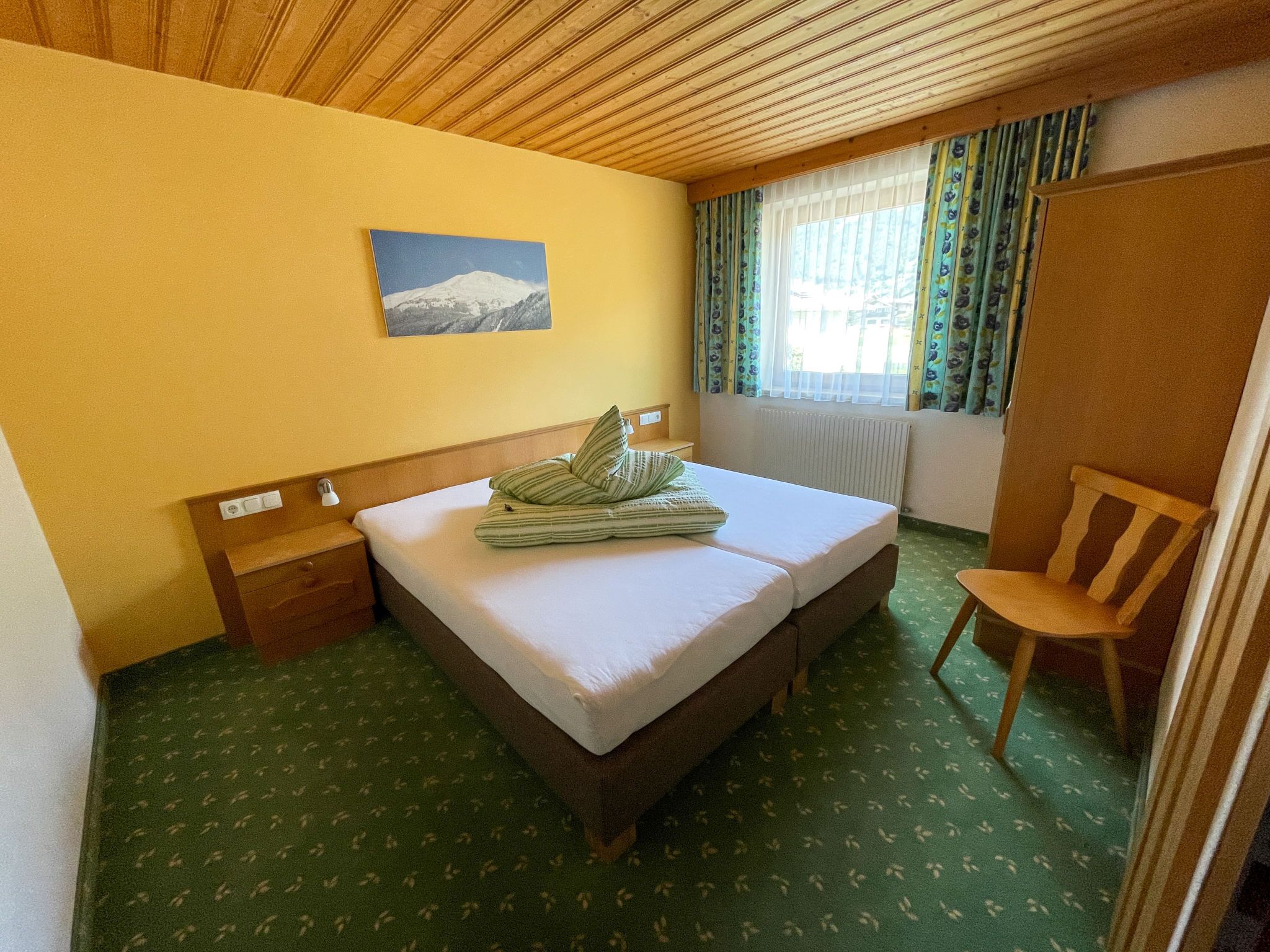Une chambre d’hôtel avec un lit, une chaise, un tableau au mur et une fenêtre avec des rideaux.