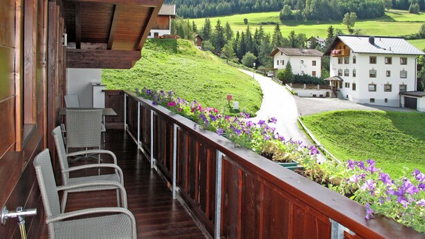 A balcony with wicker chairs, a wooden railing, and a view of a green valley with buildings and trees. - Alpenherz by Interhome (Prutz/Kaunertal)