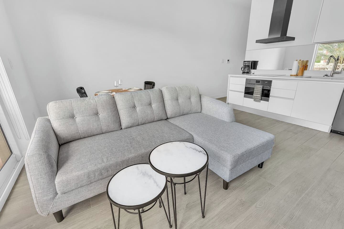 Un salon moderne avec un canapé d’angle gris, deux tables rondes surmontées de plateaux en marbre blanc, et une cuisine blanche en arrière-plan.