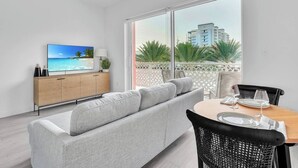 A modern living room with a grey sofa, a wooden dining table with chairs, a TV on a wooden cabinet, and a balcony with a view of palm trees and buildings. - Modern 1BR | Pool & BBQ | Steps to 7 Mile Beach (Grand Cayman)