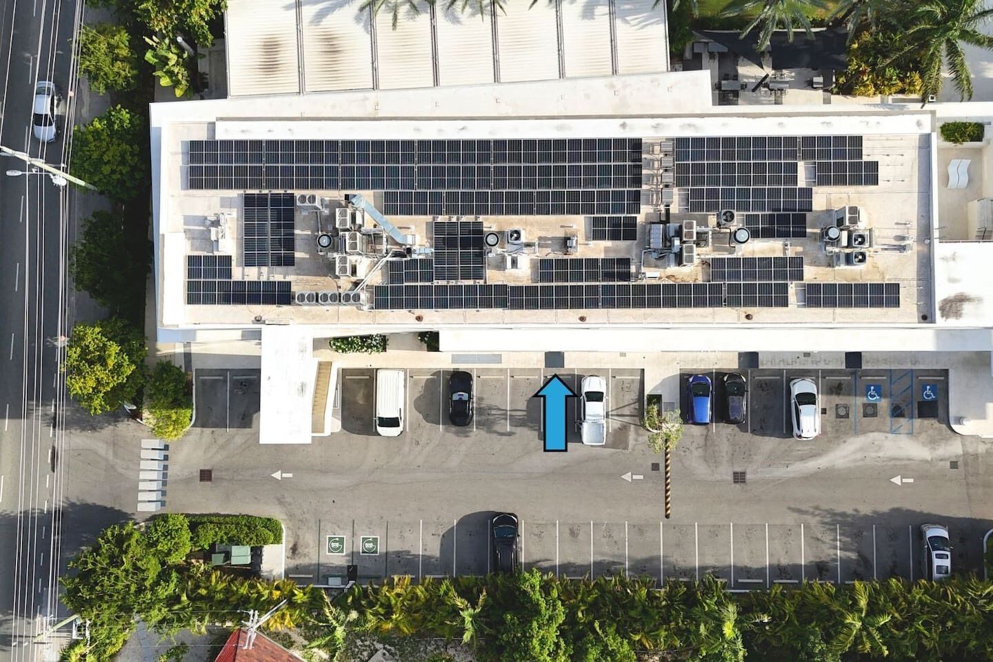 Vista aérea de un estacionamiento con múltiples filas de automóviles estacionados, una zona cubierta con paneles solares y un edificio con grandes ventanales y unidades de aire acondicionado.