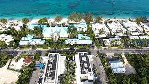 An aerial view of a coastal resort with multiple buildings, a beach, and clear blue waters. - Modern 1BR | Pool & BBQ | Steps to 7 Mile Beach (Grand Cayman)