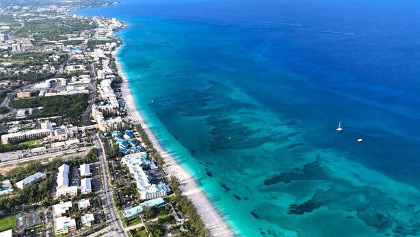 An aerial view of a coastal city with a long beachfront, clear blue waters, and a mix of residential and commercial buildings. - Modern 1BR | Pool & BBQ | Steps to 7 Mile Beach (Grand Cayman)