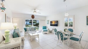 A spacious living room with a sofa, coffee table, and dining area. There is a television mounted on the wall and a ceiling fan.