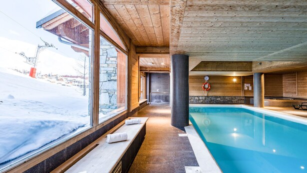 An indoor swimming pool with stone walls and wooden beams, benches along the side, and a view of a snowy outdoor area. - Les Hauts de Comborciere by Interhome (La Toussuire)