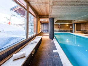 Piscina interior com paredes de pedra e vigas de madeira, bancos ao longo da borda e vista para uma área externa coberta de neve.