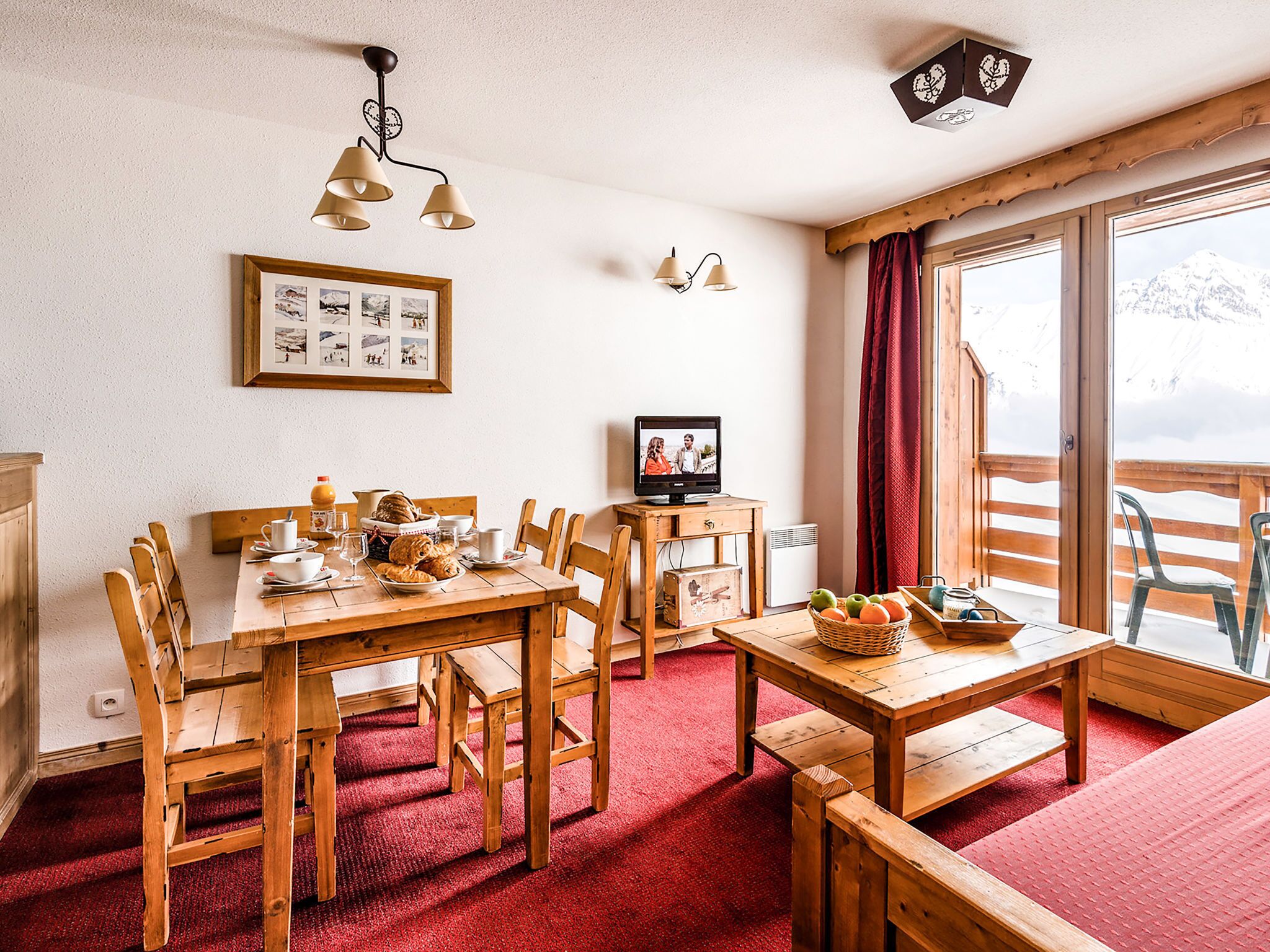 Ein gemütliches Zimmer mit einem Holzesstisch, Stühlen, einem Fernseher und einem großen Fenster mit Bergblick.
