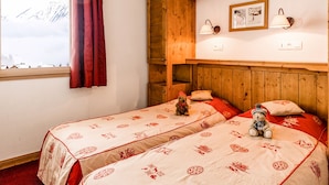 Ein gemütliches Zimmer mit zwei Betten, einem Fenster mit Blick auf schneebedeckte Berge, Holzbalken und einer kleinen Ablagefläche mit einem Teddybären.