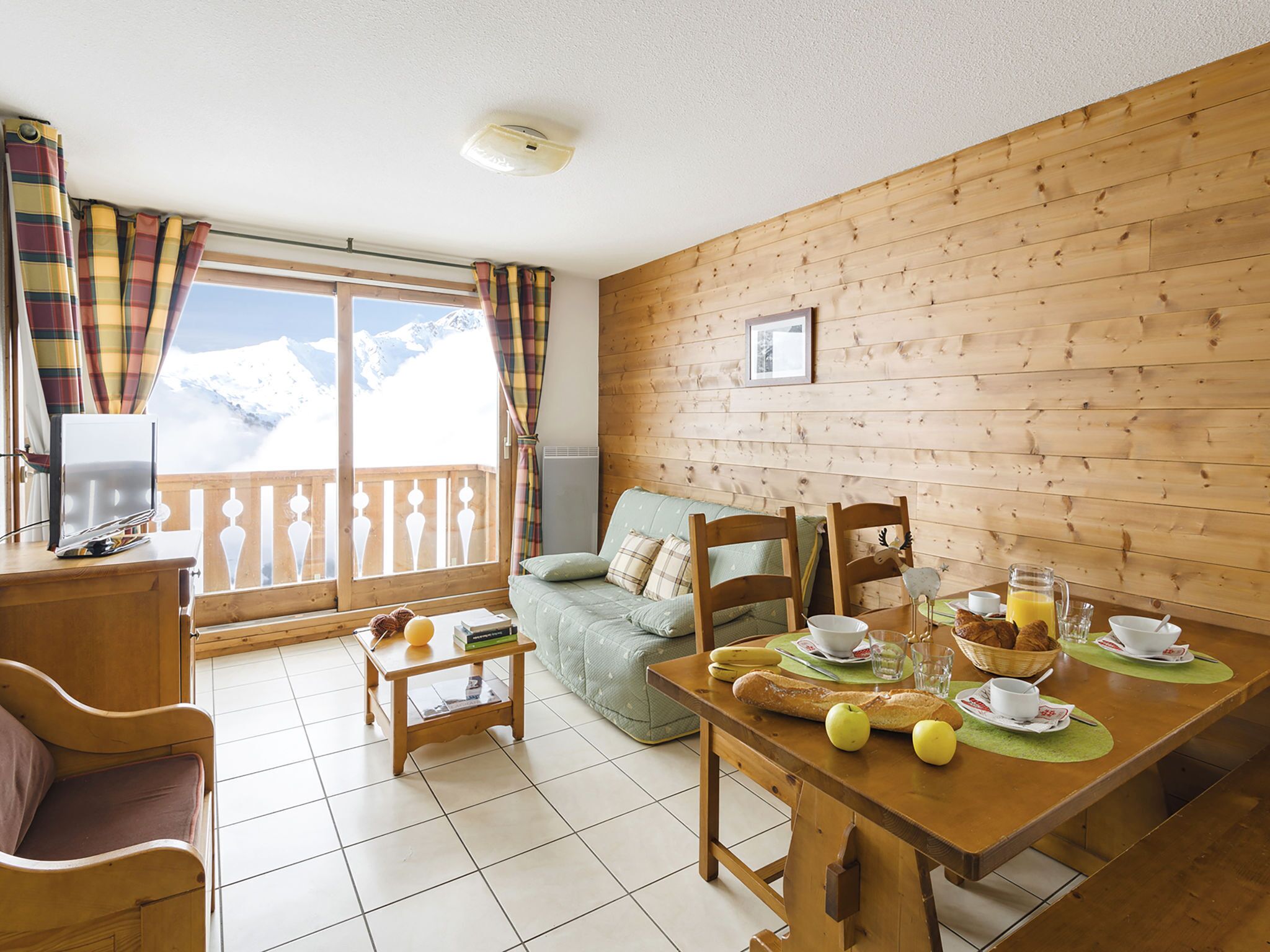 Een gezellige kamer met een houten wand, een eettafel gedekt voor het ontbijt, een bank en uitzicht op besneeuwde bergen.