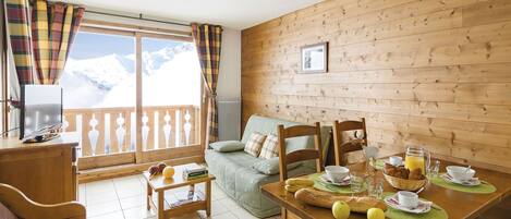 A cozy room with a wooden wall, a dining table set with breakfast, a sofa, and a view of snow-capped mountains.