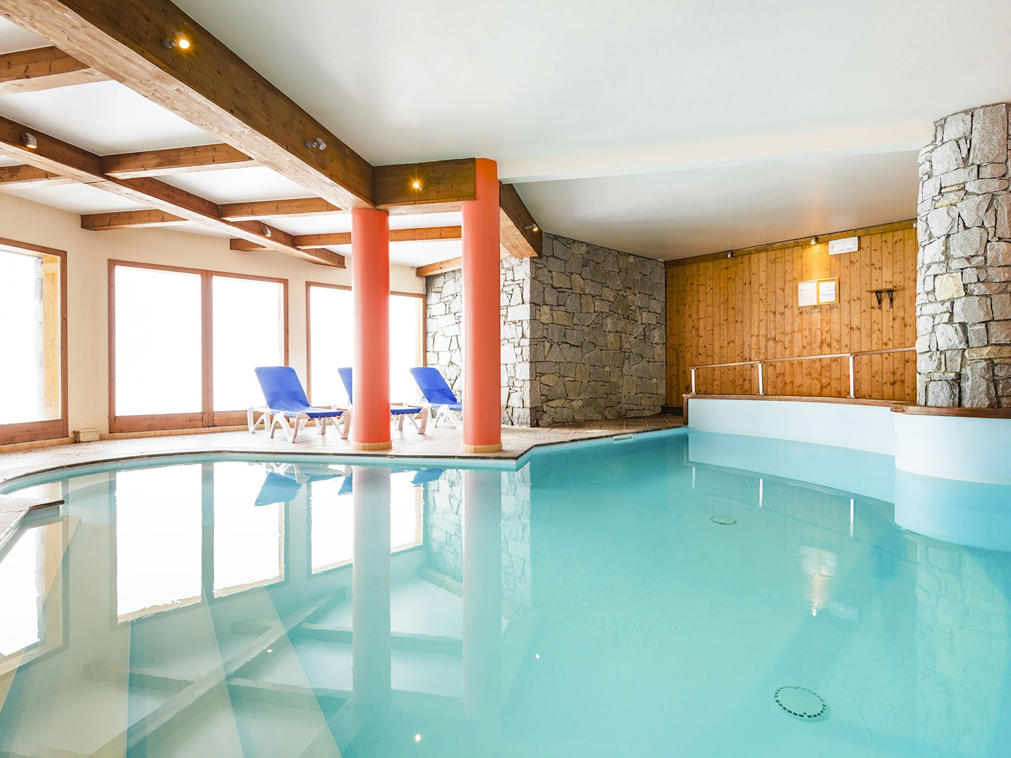 An indoor swimming pool with clear blue water, wooden beams, stone walls, and blue lounge chairs.