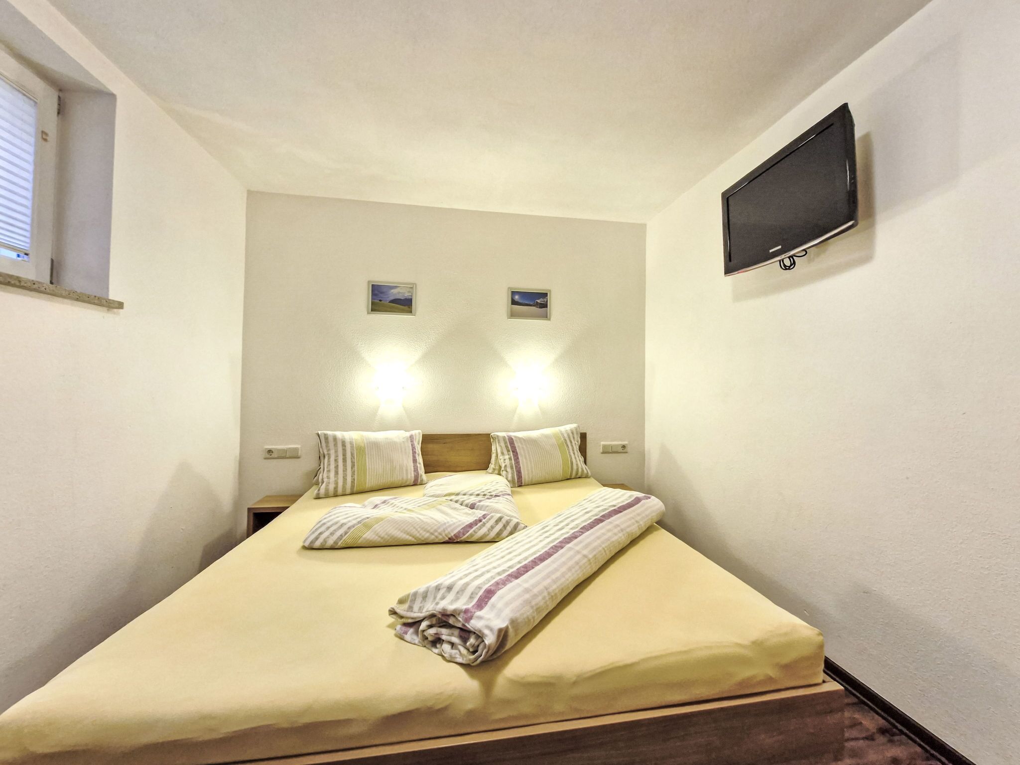 A neatly made bed with striped pillows and a rolled towel, a flat-screen TV mounted on the wall, and two small framed pictures above the bed.