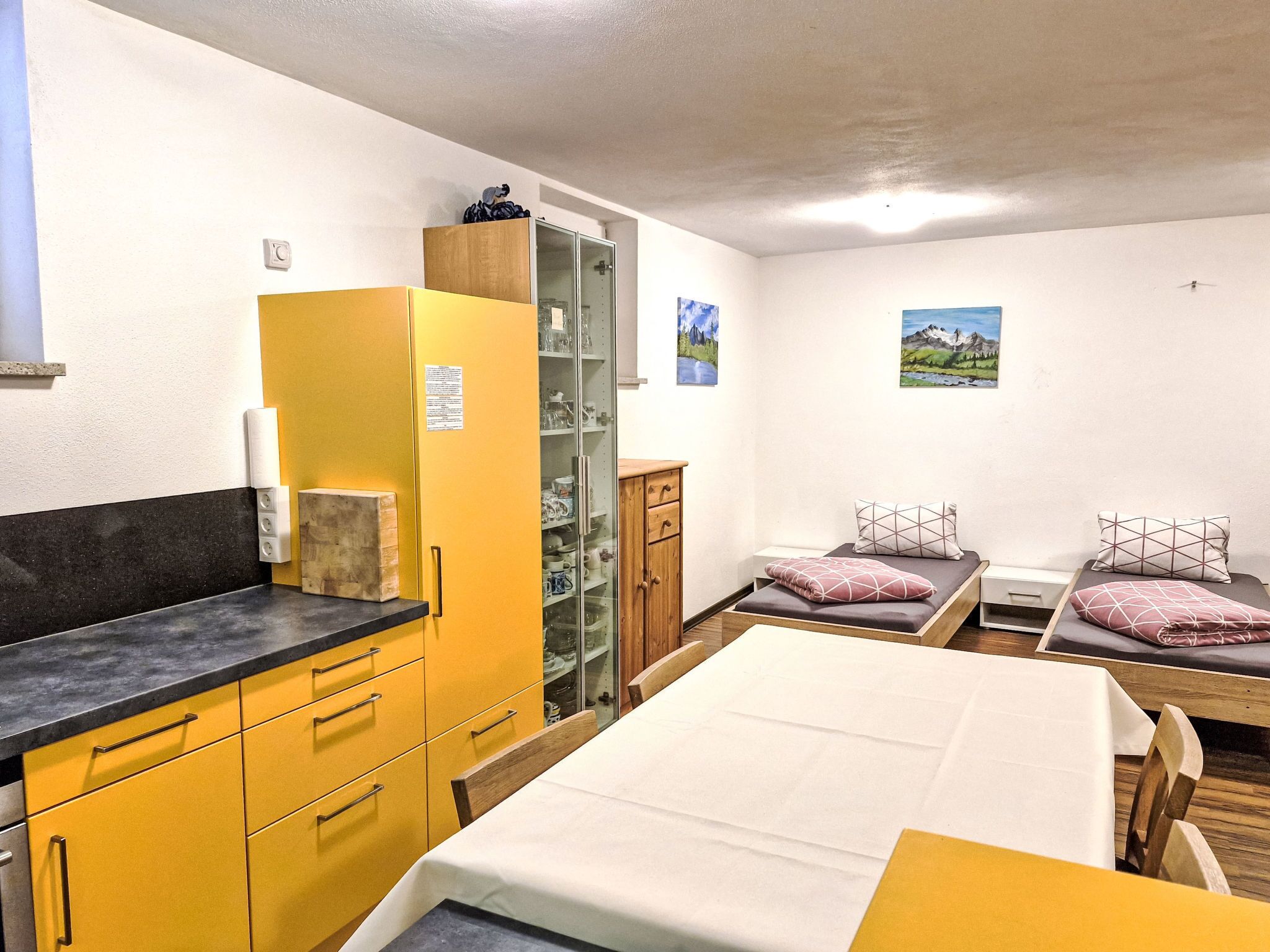 Habitación de hotel compacta con una cama, dos camas individuales, un armario de madera, una mesita pequeña y un aparador con cristal y vajilla.