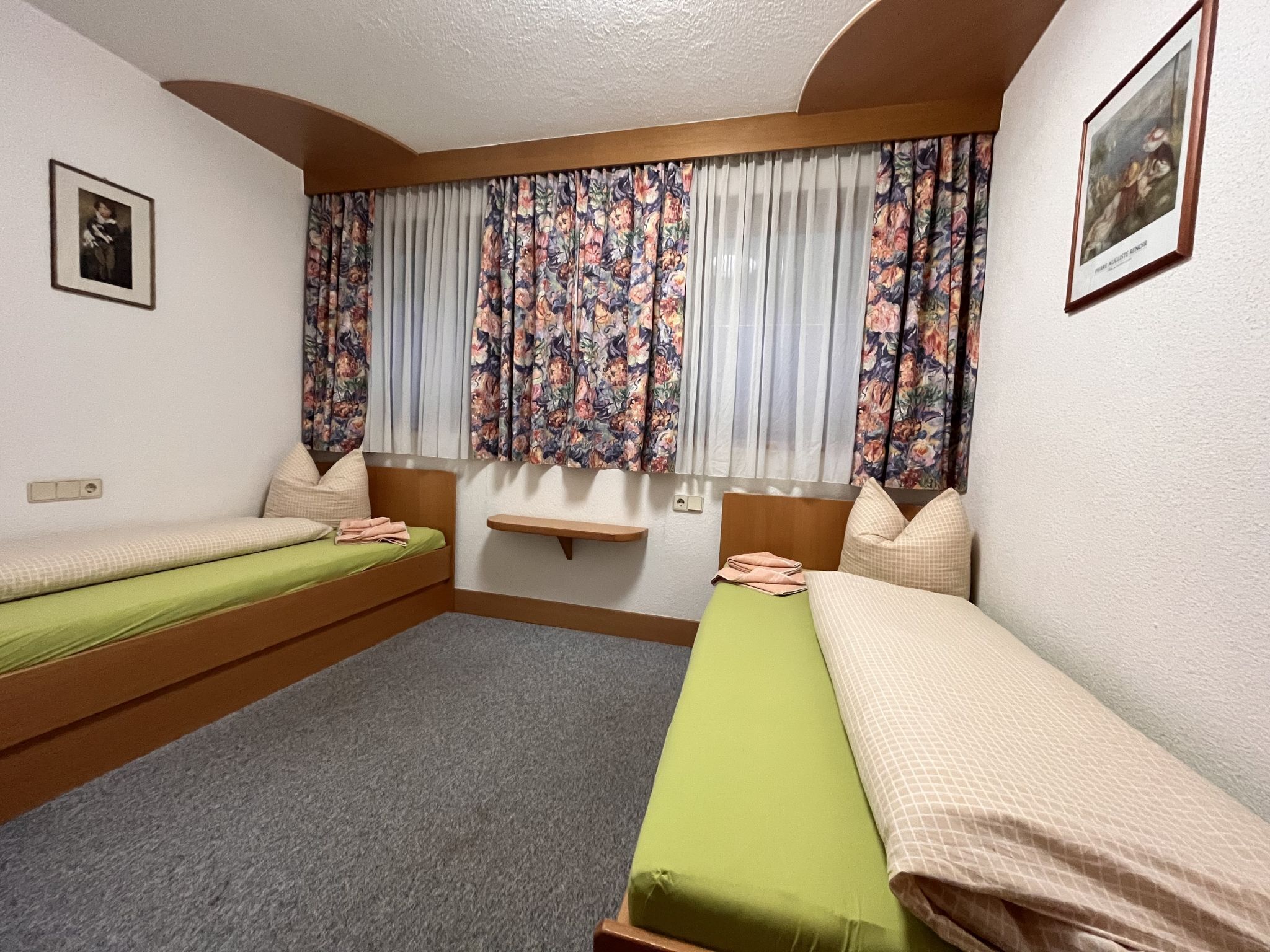 A room with two single beds, a small shelf, and a window with floral curtains.