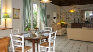 Un coin repas avec une table en bois dressée pour quatre personnes, des chaises blanches avec des assises en osier, et un mur en pierre en arrière-plan.