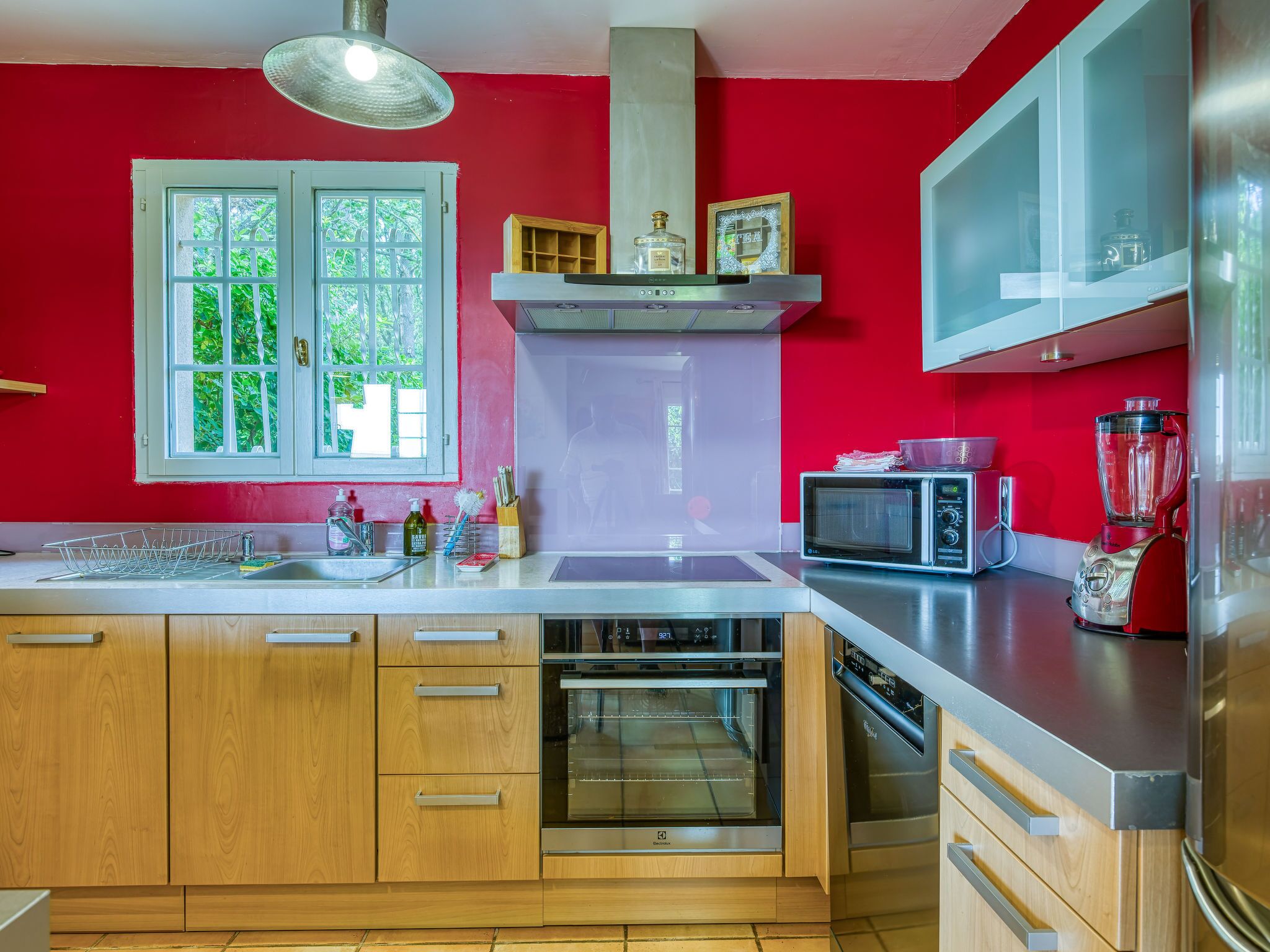A modern kitchen with red walls, a large window, stainless steel appliances, and wooden cabinets.