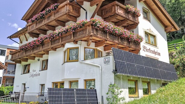 A traditional chalet-style building with wooden balconies adorned with flowers, solar panels, and a sign indicating it is a guesthouse.