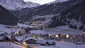 A snow-covered mountain village at dusk with illuminated buildings and a winding road.