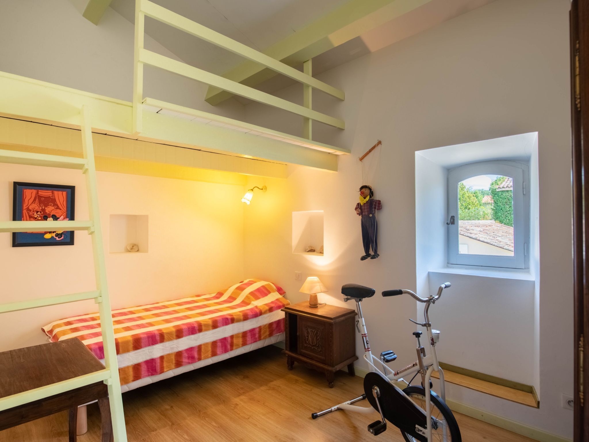 A room with a bunk bed, a desk, a lamp, a bicycle, and a window with a view of greenery.