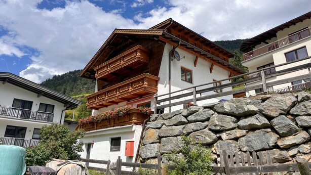 Un chalet traditionnel doté d’un balcon en bois, d’un mur en pierre et surplombant un paysage montagneux.