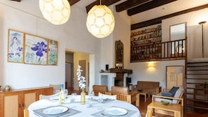 Un comedor con una mesa redonda dispuesta para cuatro personas, rodeada de sillas de madera. Se ve una estantería llena de libros al fondo, y una escalera conduce a un nivel superior.