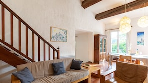 Una sala de estar acogedora con una escalera de madera, un sofá con cojines, una mesa de comedor con sillas y un ventanal con vistas a la vegetación.