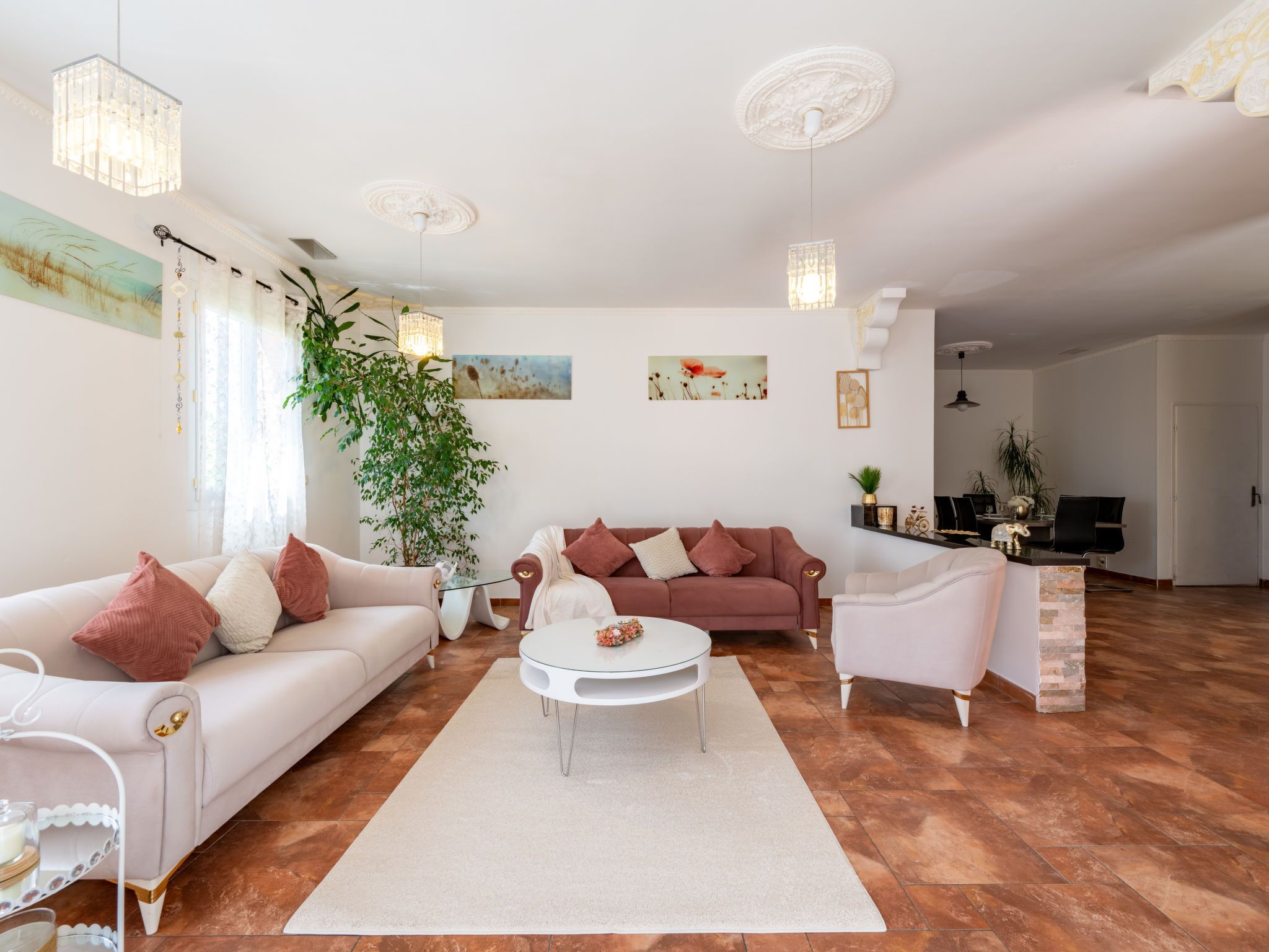 A modern living room with a white sofa, a brown sofa, a white armchair, and a round coffee table. There is a large plant, a painting on the wall, and a dining area in the background.