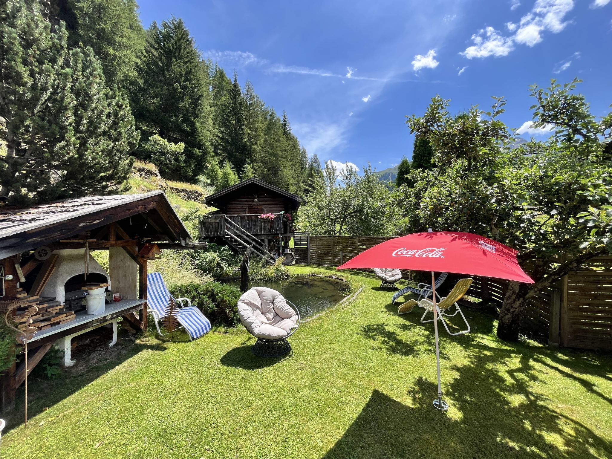 A rustic outdoor setting with a wooden cabin, a red Coca-Cola umbrella, a hammock, and a small pond.