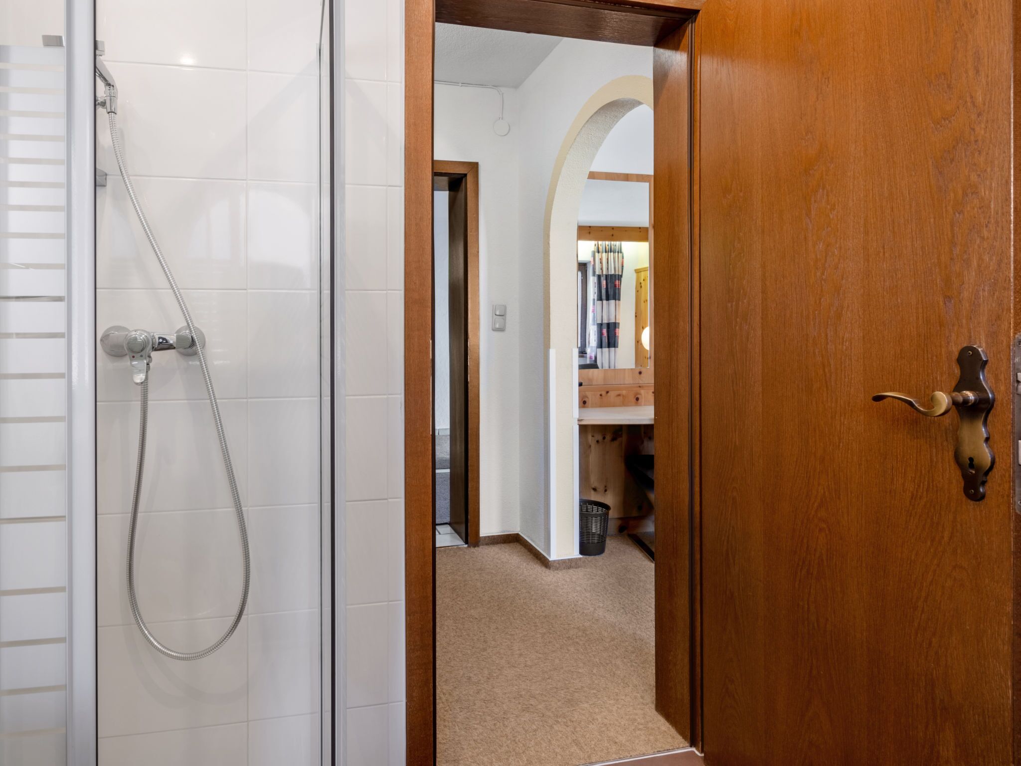 Une salle de bain avec une douche, une porte en bois et un couloir menant à une pièce équipée d’un miroir et d’un lavabo.