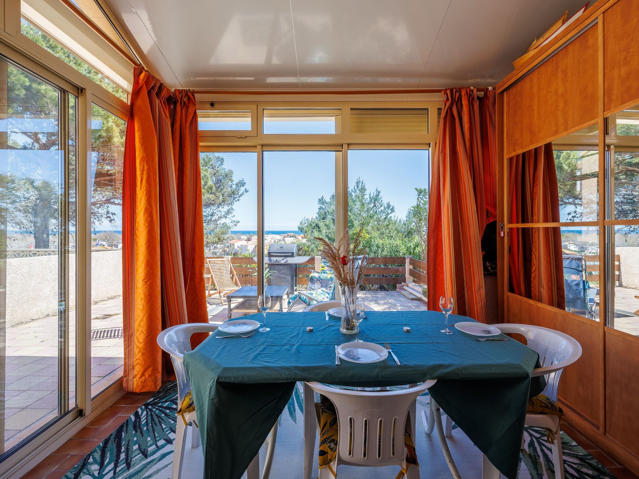 A dining area with a table set for two, large windows with curtains, and a view of the outdoors.