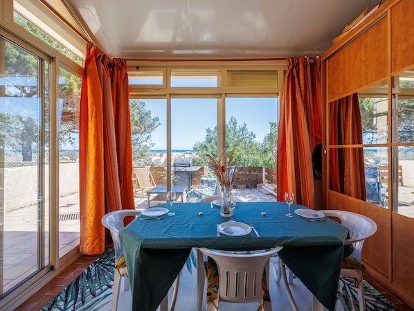 A dining area with a table set for two, large windows with curtains, and a view of the outdoors.