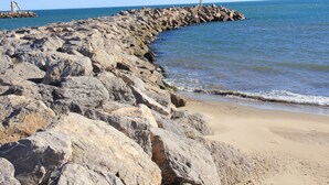 A rocky shoreline with a breakwater extending into the sea, a sandy beach, and a clear sky.