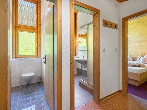 Een moderne badkamer met toilet, wastafel en douche. Een houten deur leidt naar een slaapkamer met een bed en een houten hoofdeinde.