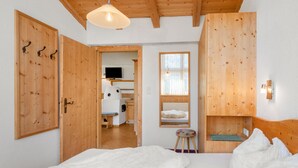 Un dormitorio con paredes de madera, ropa de cama blanca en la cama, un armario de madera, un taburete pequeño, una ventana con vistas, y un televisor en la habitación contigua.