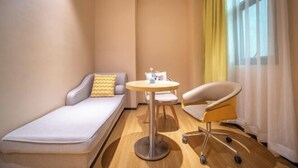 Ein kleines Hotelzimmer mit einem Einzelbett, einem runden Tisch, einem Stuhl und einem Fenster mit durchsichtigen Vorhängen.