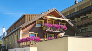 Una cabaña de madera con balcón decorado con flores púrpuras, una chimenea visible y un letrero que dice "HAUS ELISABETH".