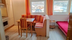 Una kitchenette compacta con estufa y horno, un rincón comedor con banco y mesa, y una cama con cubierta roja.