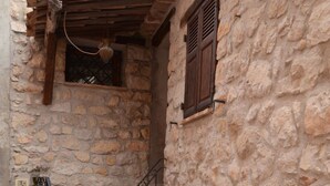 Un edificio de piedra con una escalera de madera, una pequeña ventana con contraventanas y una planta en maceta con flores rojas.