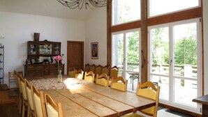 Ein Essbereich mit einem großen Holztisch, gelb gepolsterten Stühlen, einer Holzkommode und Blick durch große Fenster ins Freie.