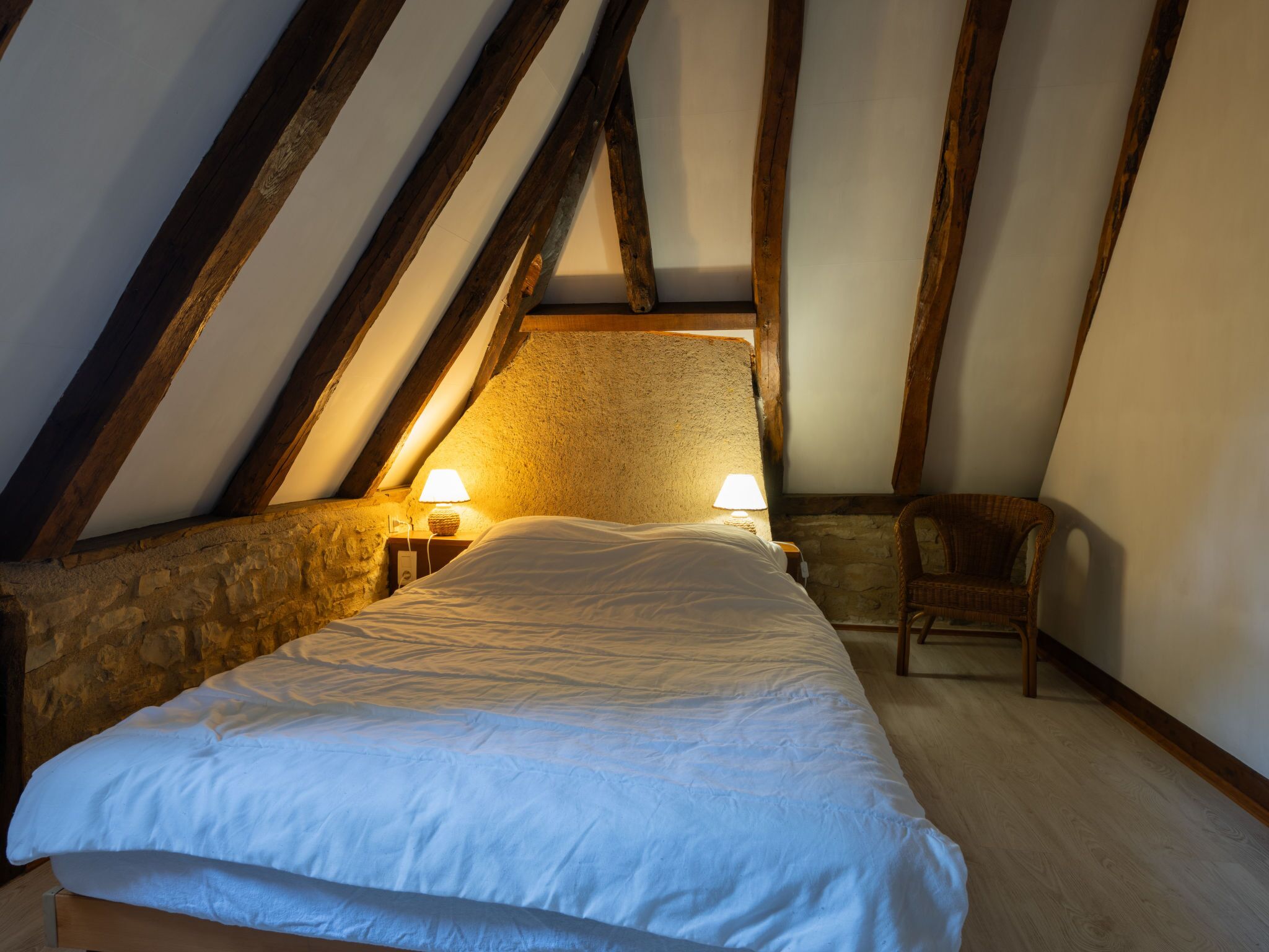 Un dormitorio con techo inclinado, vigas de madera, paredes de piedra, cama con ropa blanca, dos lámparas de noche y una silla de mimbre.
