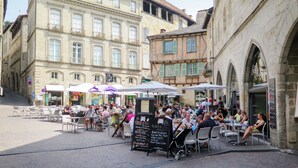 Un café al aire libre concurrido, con mesas y sombrillas, rodeado de edificios históricos con fachadas de piedra y ventanas arqueadas.
