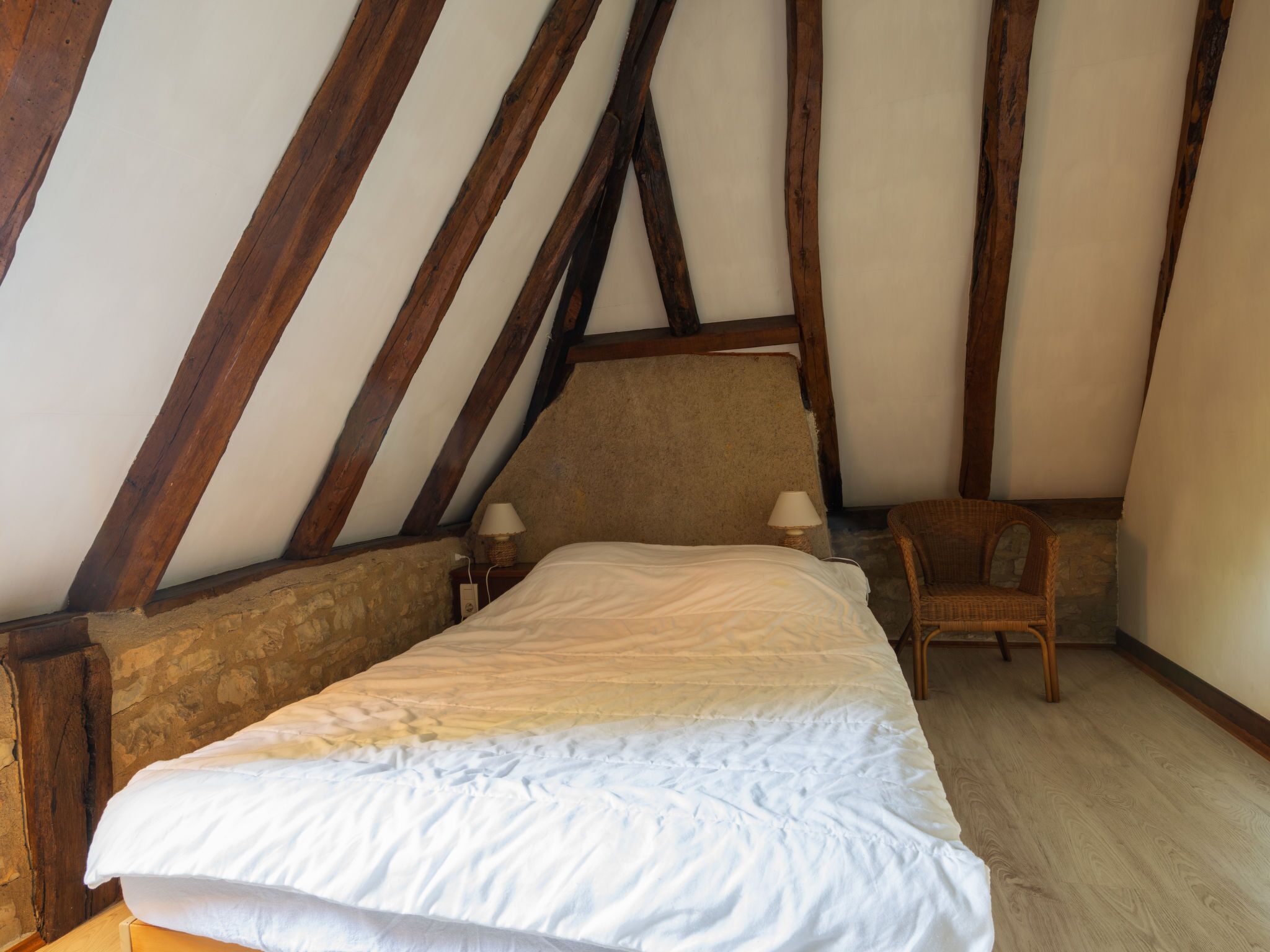 Ein Schlafzimmer mit Holzbalkendecke, einem Bett mit weißer Bettwäsche, zwei Nachttischlampen und einem Korbstuhl.