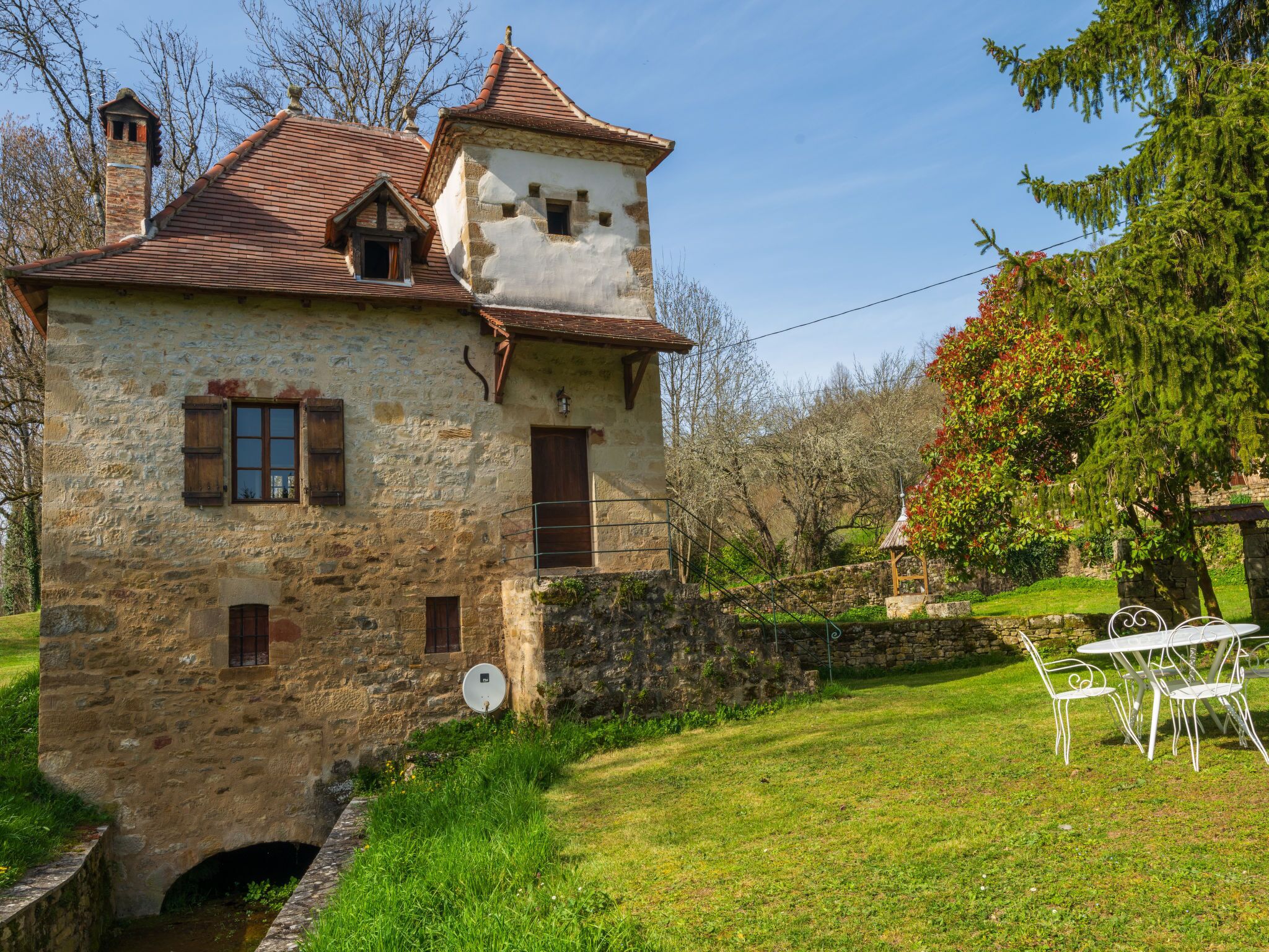 Ein Steinhaus mit Ziegeldach, einem kleinen Turm und einer Satellitenschüssel. Ein Rasenstück mit einem weißen Metalltisch und Stühlen.