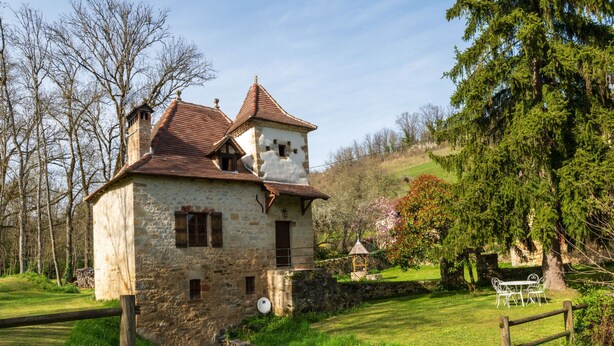 Un edificio de piedra con tejido rojo, una pequeña torre y un puente de piedra que conduce a un área verde con un pabellón y sillas de metal blanco.